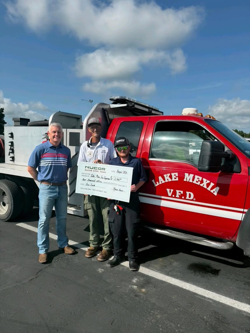 Three adults holding a ceremonial presentation check beside a red Lake Mexia VFD vehicle.